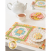 A shot of the placemat, staged atop a white table with a white teapot, tea cup, and plate of decorated cookies.