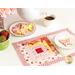 One of the June placemats, colored in bright pink, red, white, and yellow, staged with plates of iced butterfly cookies and a teapot pouring a cup of tea.