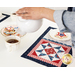 A model pours tea into a white cup over a July placemat with a white plate holding a coordinating iced cookie.