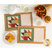 A top down shot of the two completed mats, demonstrating the warm autumn fabrics and creative piecing. Coordinating decor and a plate of cookies frames the mats on a white table.