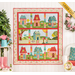 The completed Welcome Home In Spring quilt in bright, cheerful colors, hung on a paneled wall beside pink rainboots, a watering can planter with pink flowers, and a wreath of pink roses. A yellow banner in the upper right hand corner reads 