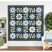 The Edelweiss quilt, completed in navy blue, powder blue, green, and white, hung on a white paneled wall and staged beside coordinating plants, furniture, and decor.