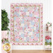 Photo of a finished pink, blue, white, and green floral quilt hanging on a white wall, with houseplants, pink flowers, and a white stool and shelf on either side
