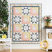 The completed Patchwork Sentiments quilt in pink, yellow, white, and navy blue, hung on a white paneled wall and staged with coordinating housewares and flowers.