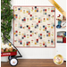 A playful beige quilt hung on a white wall next to a wagon full of flowers