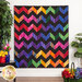 The completed Charming Chevrons Quilt, hung on a white paneled wall and staged with coordinating flowers and houseplants. A basket of rainbow florals can be seen in the lower right corner.