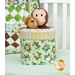A head on shot of the completed Fabric Basket in Wee Safari prints, staged on a white bed with green dressings. The basket holds a stuffed monkey and stuffed lion toy. In the background, the Little Pathways Quilt can be seen draped over the headboard of the bed.