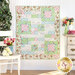 The completed Comfort of Psalms quilt in Serene Garden, a palette of green, blue, pink, and cream floral prints. The quilt is hung on a white paneled wall and staged with coordinating furniture and decor.