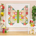 The completed Butterfly Patch Quilt in white and bright colors from Moda's Strawberry Lemonade collection; it is hung on a light blue paneled wall and staged with coordinating decor and flowers.