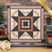 A wide shot of the completed Prairie Star Quilt in rich earth tones from the Chickadee Landing collection. The quilt is hung on a rustic wood paneled wall and staged with coordinating furniture and decor. A yellow banner in the upper right hand corner reads 