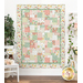 A finished pink, white, and green floral quilt hanging on a white wall, with houseplants, flowers, and a white chair and shelf on either side.