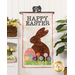 The completed Happy Easter Door Banner, hung on a white paneled wall and staged with coordinating foliage, decor, and furniture.