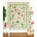 A finished pink, white, and green floral quilt hanging on a white wall, with houseplants, flowers, and a white chair and trellis on either side.