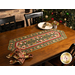 The Completed Up On The Housetop Table Runner, colored in green and red, staged on a brown table with coordinating Up On The Housetop napkins and Christmas decor.