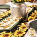 A close up of the table runner on a white table showing detail on the sunflower and butterfly fabrics and staged with coordinating plates, glassware, and a tin holding decorative sunflowers.