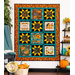 The finished Gather Together panel quilt in warm orange and teal colors, hung on a white paneled wall with a shelf on the right covered in sunflower and fall-themed decor. On the left, a leafy tree sits behind a knit blanket that is thrown over a chair.