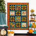 The finished Gather Together panel quilt in warm orange and teal colors, hung on a white paneled wall with a shelf on the right covered in sunflower and fall-themed decor. On the left, a leafy tree sits behind a knit blanket that is thrown over a chair.