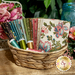 A wicker basket filled with colorful fabric pieces and spools of thread, surrounded by flowers.