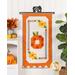 The completed October Door Banner, a lovely pumpkin with patchwork floral accents colored in bright orange, white, and yellow, hung on a white paneled wall and staged with coordinating decor.