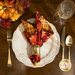 A top-down shot of a beautifully set dinner table featuring a plate with the cloth napkin atop beside utensils, a glass, and decor; the leaf print faces outward.