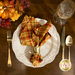 A top-down shot of a beautifully set dinner table featuring a plate with the cloth napkin atop beside utensils, a glass, and decor; the plaid print faces outward.