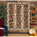 The completed Pumpkin Lane quilt, colored in shades of tan, red, orange, green, and navy; two rows of laser cut applique pumpkins run between three rows of on point patchwork. It is hung on a brown wood paneled wall and staged next beside coordinating furniture and flowers.