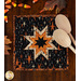 A black square quilted pot holder with an orange star pattern, accompanied by wooden cooking spoons resting on top while coordinating seasonal decor peeks into the top and left sides of the frame.