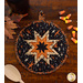 A black round quilted pot holder with an orange star pattern, accompanied by a wooden cooking spoon resting on the table next to the hot pad while coordinating seasonal decor peeks into the right and left sides of the frame.