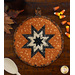 An orange round quilted pot holder with a black and cream star pattern, accompanied by a wooden cooking spoon resting on the table next to the hot pad while coordinating seasonal decor peeks into the right and left sides of the frame.