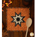 An orange square quilted pot holder with a black and cream star pattern, accompanied by a wooden cooking spoon resting on top while coordinating seasonal decor peeks into the top eft side of the frame.