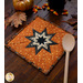 A square quilted pot holder featuring a star design in black and cream on an orange background, with a wooden spoon beside it. There is coordinating seasonal decor in the background as the hot pad sit on a dark wooden counter.