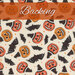 A swatch of cream fabric with tossed pumpkins wearing witch hats, bats, and sparkles. A burnt orange banner reads 