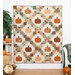 The completed Pumpkin Patches Quilt, colored in elegant and muted colors like cream, burnt orange, and eucalyptus, hung on a white paneled wall and staged with pumpkins and coordinating decor.