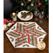 The completed 60 Degree Diamond Table Topper, colored in red, green, and white fabrics from the A Christmas Carol collection by Moda Fabrics, staged on a rustic wood table with coordinating decor. A colorful Christmas tree peeks into frame from above.