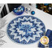 A shot of the completed Point of View Folded Star Table Topper Kit in fabrics from the Holiday Flourish - Festive Finery collection, a series of wintry blue metallic prints. It is staged on a white table with coordinating blue and winter decor.