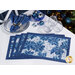 The four completed self binding placemats, colored in wintry blue and white prints from the Holiday Flourish Festive Finery collection, staged on a white table with coordinating glasses, napkins, table settings, and decor.