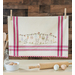 A vintage style dish towel with hand embroidered bunnies and Easter motifs hanging from a clothesline over a kitchen counter with a rolling pin, kitchen utensils, and eggs.