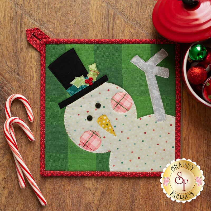 Hot Pad with a laser cut applique snowman waving laid flat on a wood table