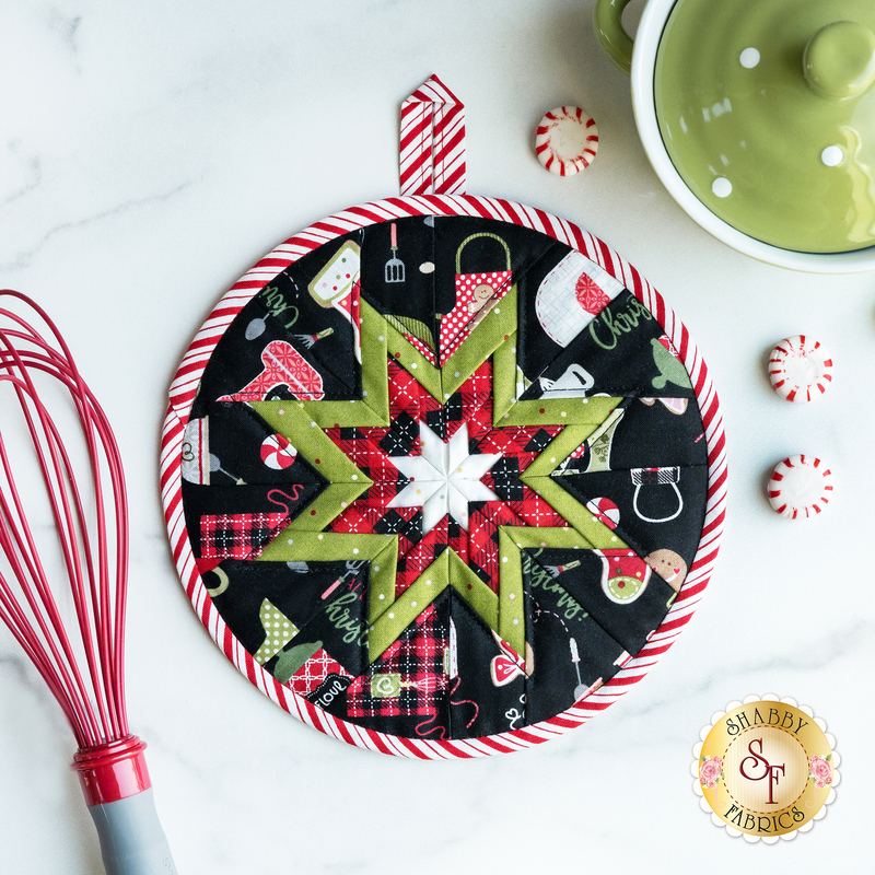 Black, red, and green Christmas themed hot pad on a white table with peppermint candies and a red whisk and green pot