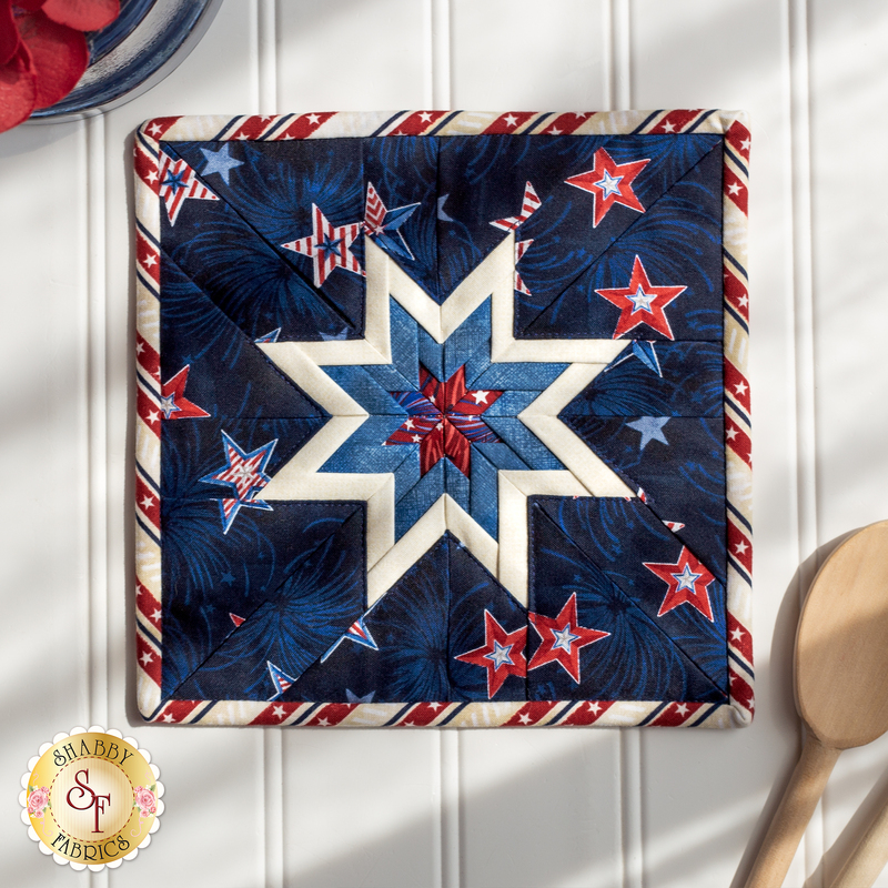 Red, white, and blue folded star squared hot pad on a white paneled background with a wooden spoon nearby