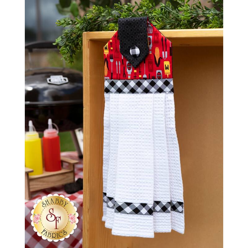 Red and black Peace, Love & BBQ Hanging Towel hung outdoors in front of a BBQ and picnic table