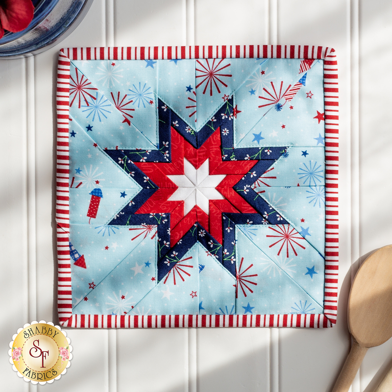 The light blue Red, White, & Bloom Folded Star Squared Hot Pad on white wood paneling with a wooden spoon nearby