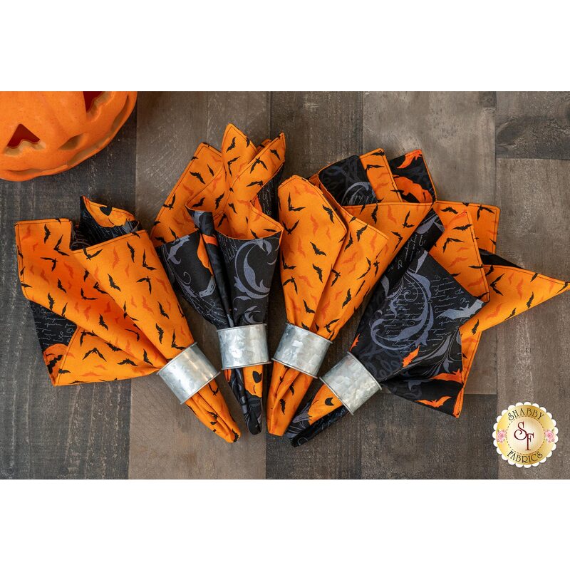 Four orange and black Halloween themed cloth napkins on a wood table next to jack-o-lantern.