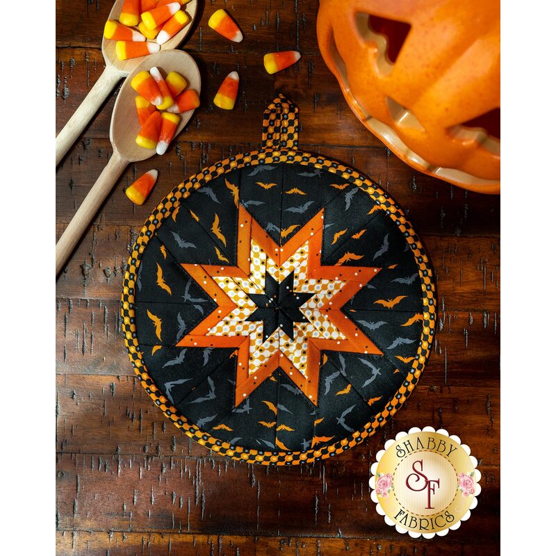 Black and orange Halloween themed hot pad on wood table next to spoons, candy corn, and a jack-o-lantern.