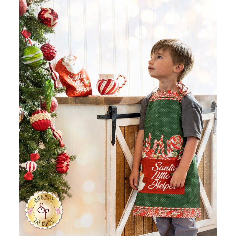 Photo of a child wearing the Peppermint Candy apron looking up at a Christmas tree, standing next to a counter with the Santa's Little Helper bag and a peppermint candy mug