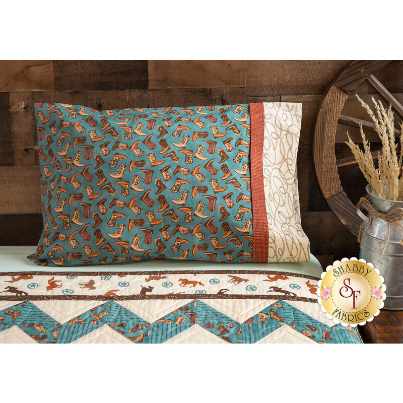 Teal colored pillowcase featuring cowboy boots resting on bed with matching cowboy themed quilt.