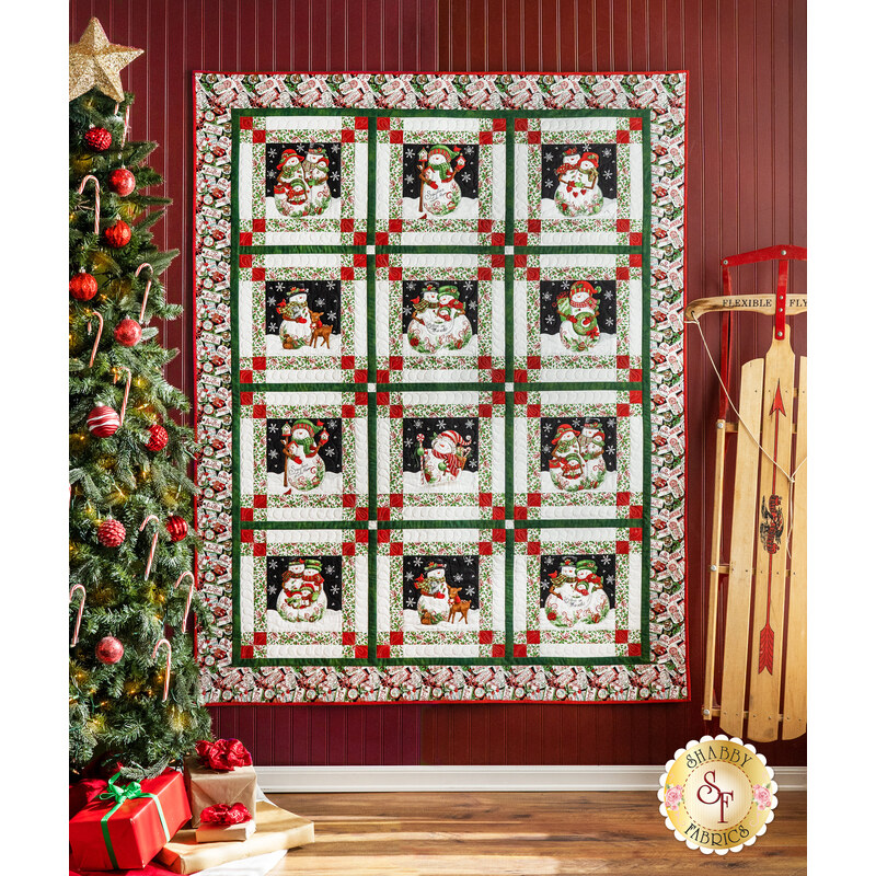12 block quilt featuring snowman illustrations and holiday colors, hanging on a red wall next to a Christmas Tree.