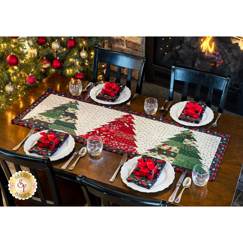 White table runner featuring 3 red and green Christmas trees on white as part of a table setting including plates and silverware.