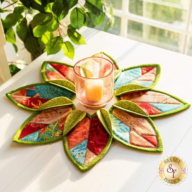Leaf shaped table topper with central opening containing a candle. Topper is made of red, green, and teal floral fabrics.