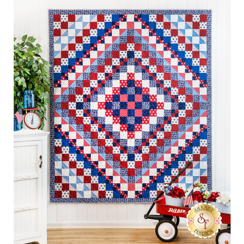 Red, white, and blue quilt of squares in diamond pattern with patriotic themed fabrics hanging from a white panel wall.